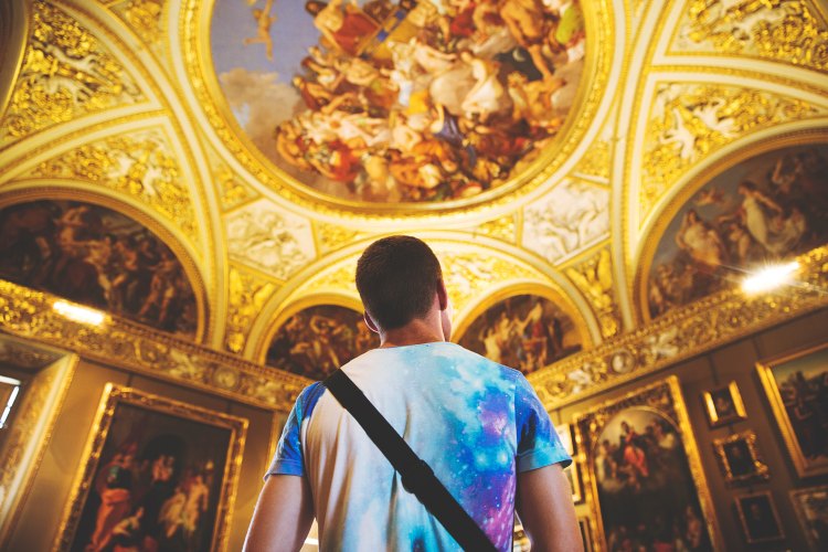 A young man in a colorful galactic t-shirt gazes up at an ornate gilt and fresco ceiling depicting paradise, with Jesus Christ seated on a throne surrounded by a joyful crowd.